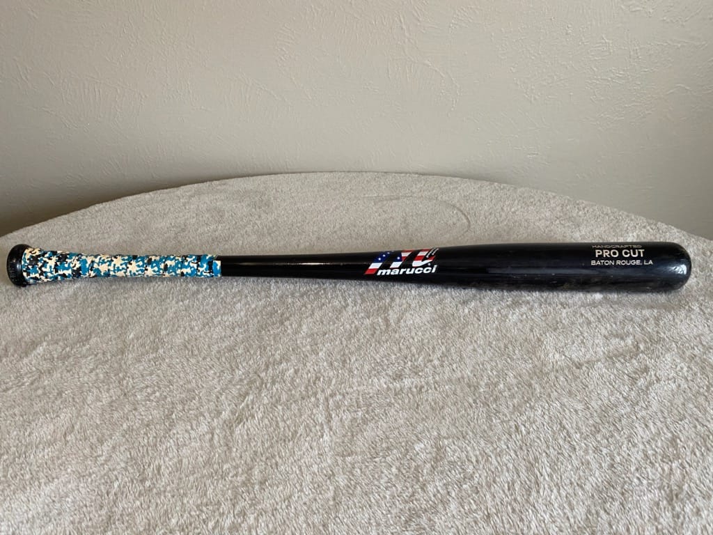 Black Marucci baseball bat with blue and white grip laying flat on a white blanket