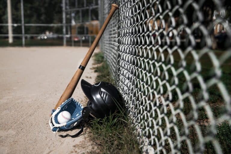 A baseball bat resting against a fence next to a batting helmet and a youth baseball glove with a baseball inside