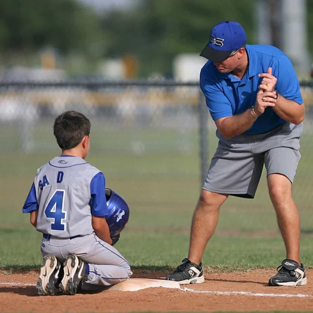 A youth baseball player kneeling on a base with two knees while a coach demonstrates a swing