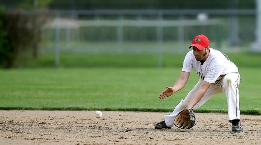An infielder wearing a white uniform and red hat gets into position to field a ground ball