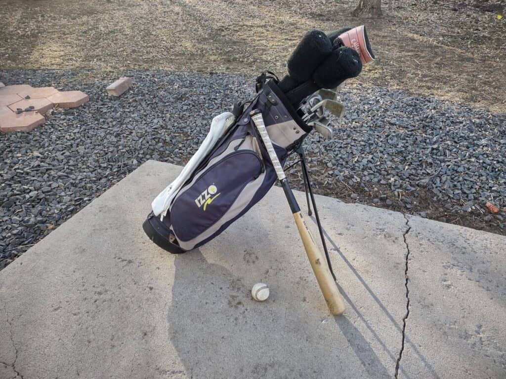 A baseball bat resting on a set of golf clubs with a baseball laying nearby