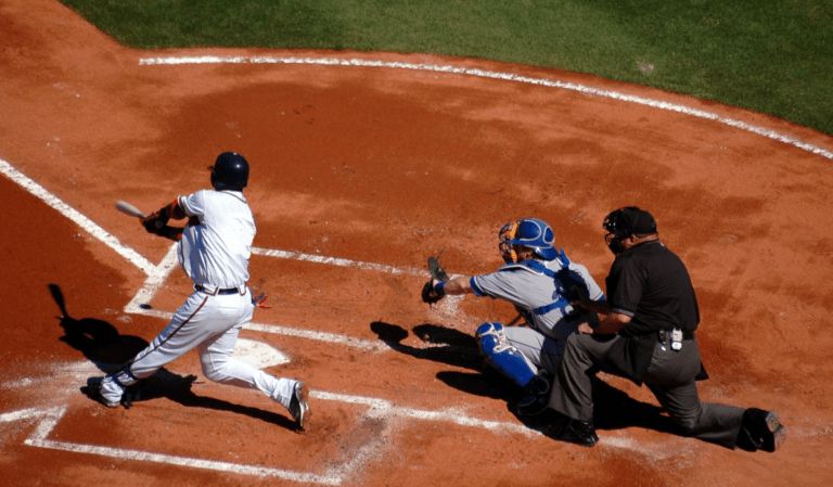 A right-handed hitter in a white uniform in the batter's box swinging at a pitch while the catcher reaches to catch the ball