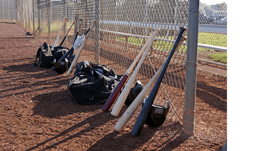 Baseball bats, batting helmets, and duffle bags resting along the fence by the dugout