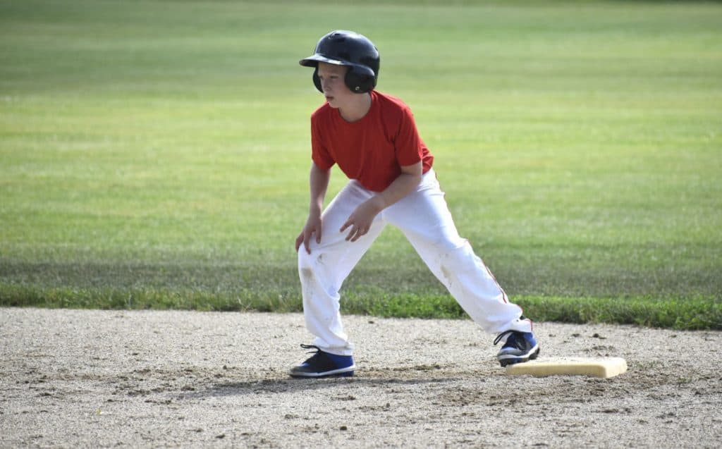 Youth baserunner in a red shirt is standing on second base and waiting for the pitch to be delivered