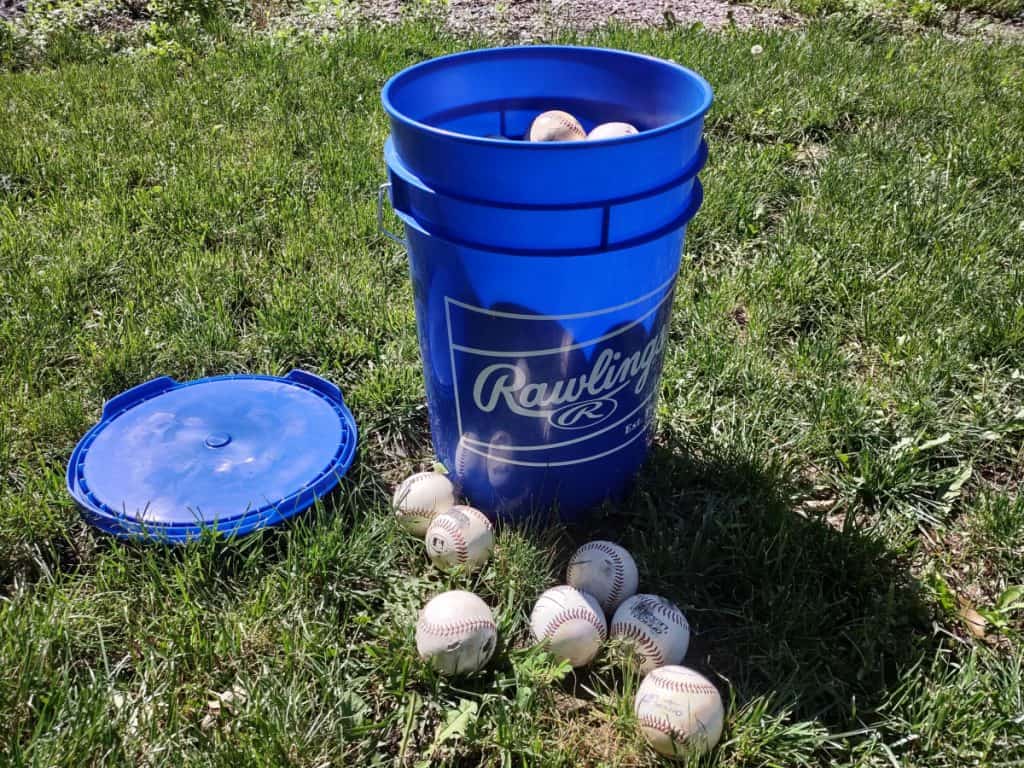 A 6-gallon bucket of baseballs in the grass with the lid and extra baseballs laying around the bucket