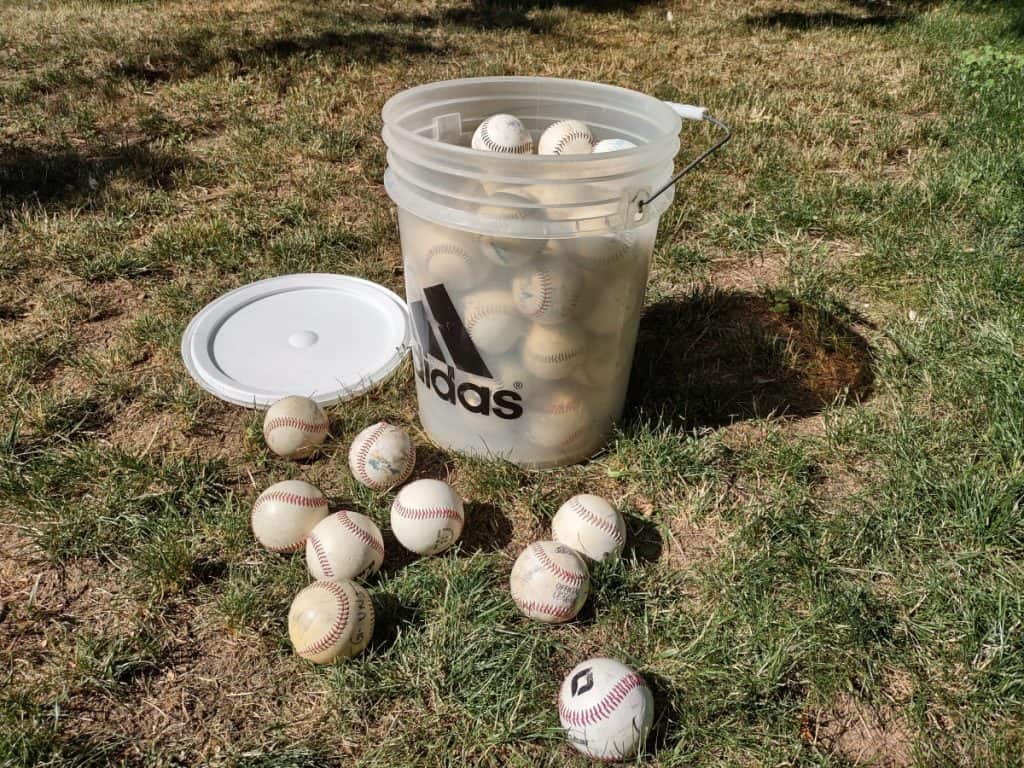 A 5-gallon plastic adidas bucket filled with baseballs with the lid and extra baseballs laying on the ground