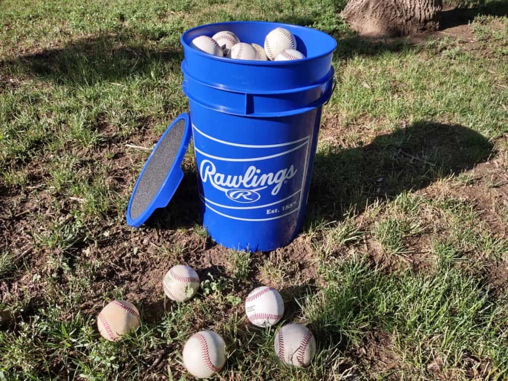 A full 6-gallon bucket of baseballs on the grass with the lid resting against the bucket and five extra baseballs laying around the bucket