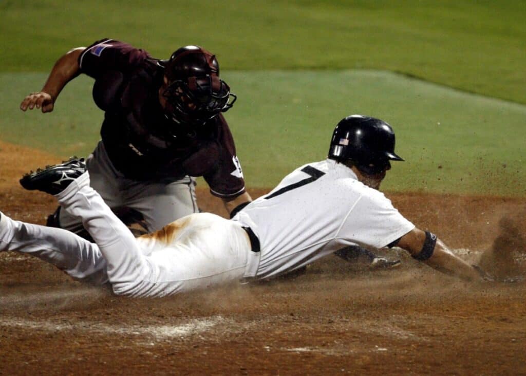 Baseball player in white uniform slides headfirst into home plate while the catcher in a maroon uniform applies the tag