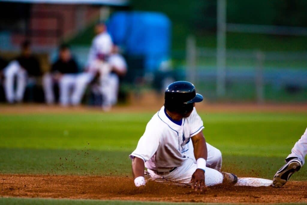 Baseball player in an all-white uniform slides feet-first into the base with the left foot of the defender also touching the base