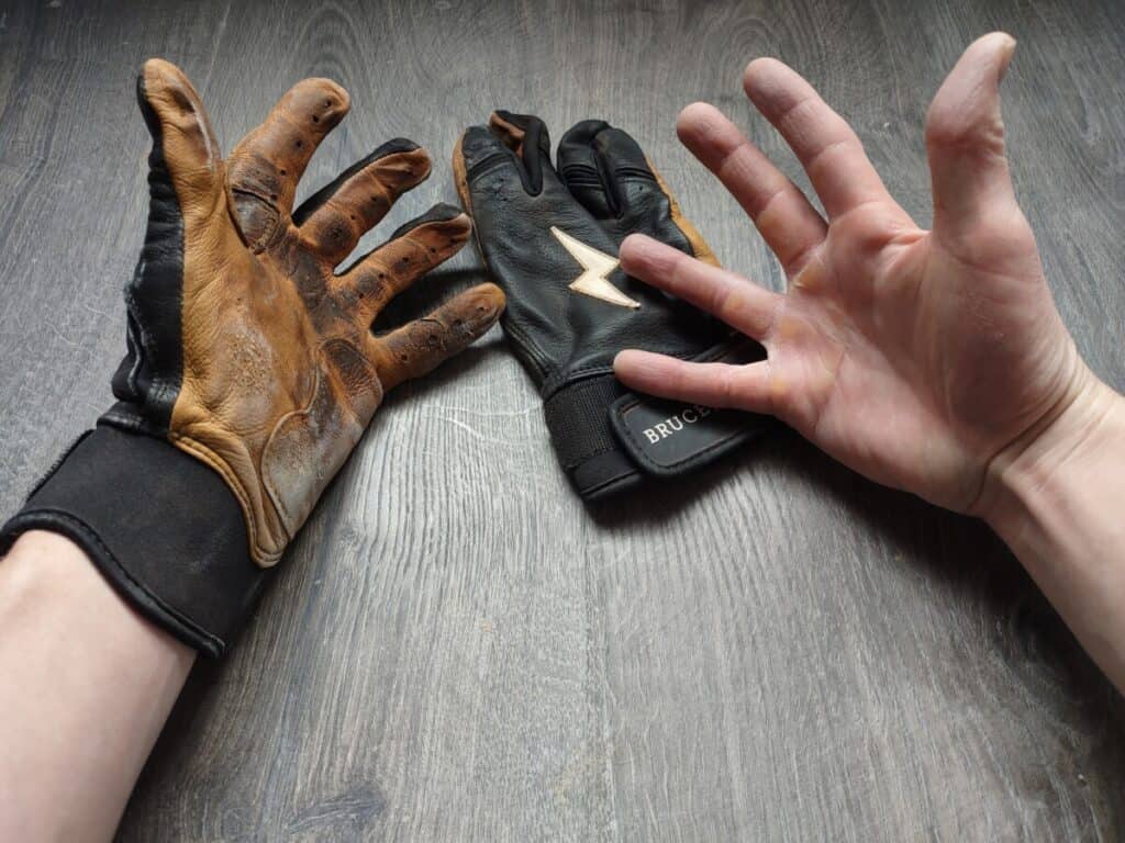 Left hand wearing a Bruce Bolt batting glove while the other Bruce Bolt batting glove lays on a wood floor next to the right hand