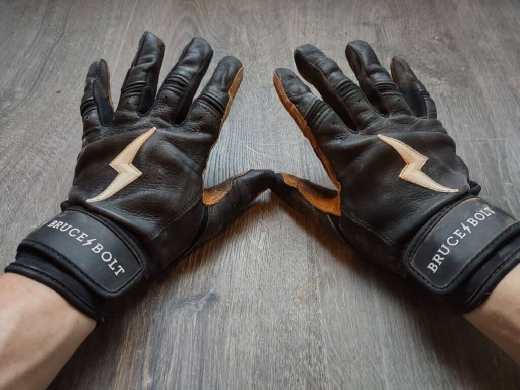 Two hands wearing Bruce Bolt batting gloves resting on a wood floor