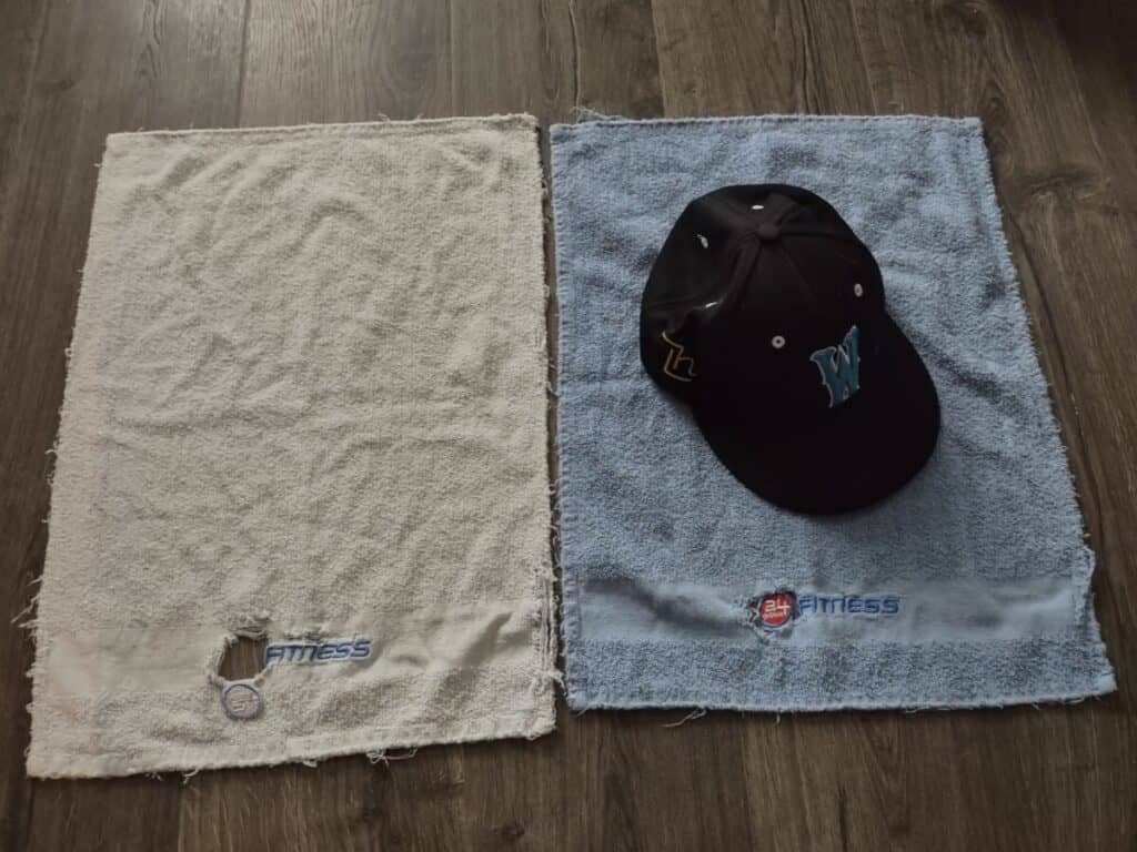 Two old gym towels that are used for the baseball towel drill are laying a floor. There's a baseball hat laying on top of the blue gym towel.