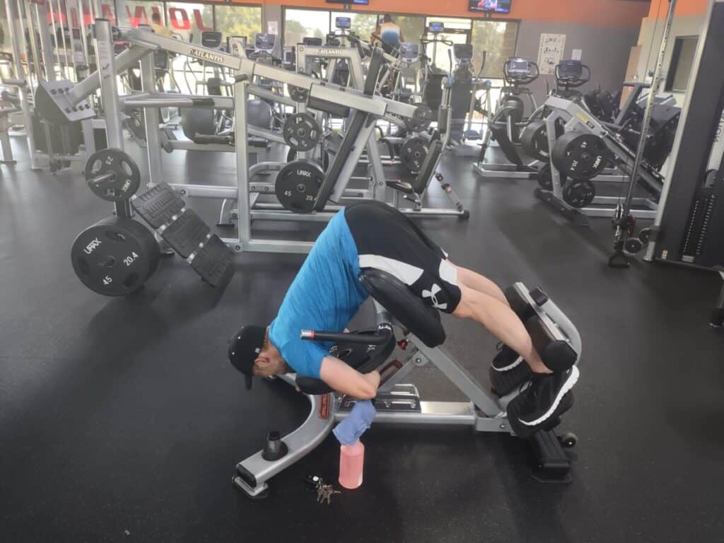 Steve Nelson in the starting position for the back extension exercise. Legs are secured by leg pads, thighs are resting on the thigh pads, and player is bent over and holding a weight