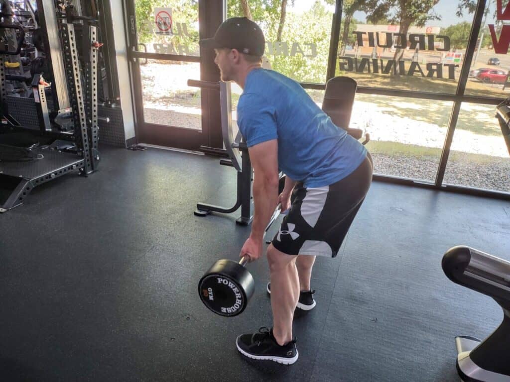 Demonstrating the starting position of the bent over low row exercise with a straight bar. Player is using a reverse grip with his palms facing outward, his back is at a 45-degree angle, and the bar is hanging just below his knees.
