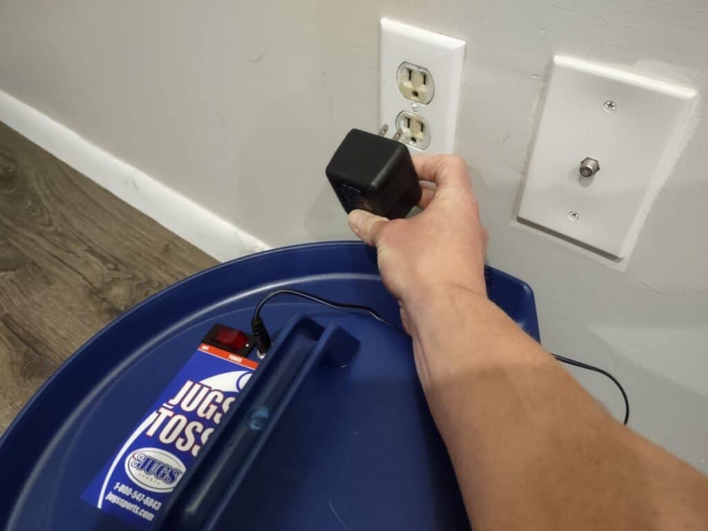 Inserting the plug into a wall outlet to charge a Jugs Soft Toss machine