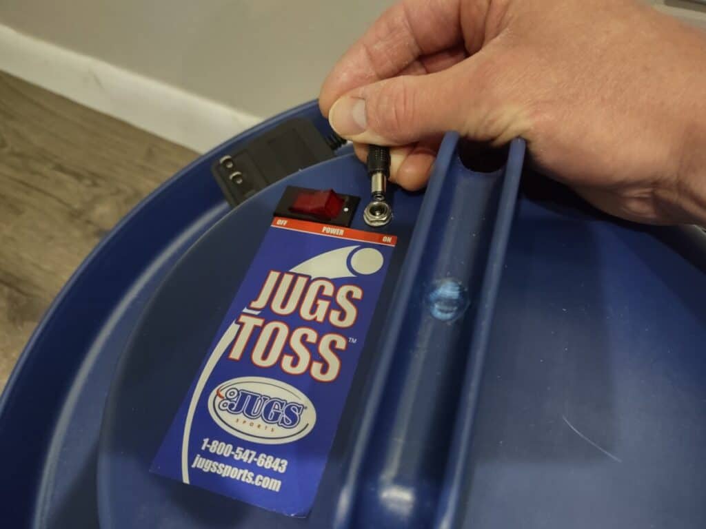 Plugging the small, round end of the charging cord into the top of a Jugs Soft Toss machine