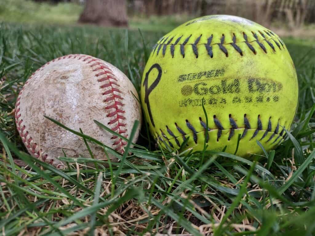 Used leather baseball laying in the grass next to a used yellow softball