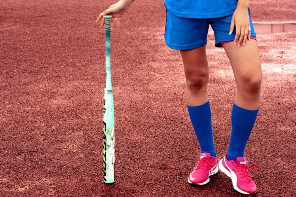 Softball player wearing blue shorts, blue socks, and red shoes is holding a softball bat with the barrel of the bat touching the ground