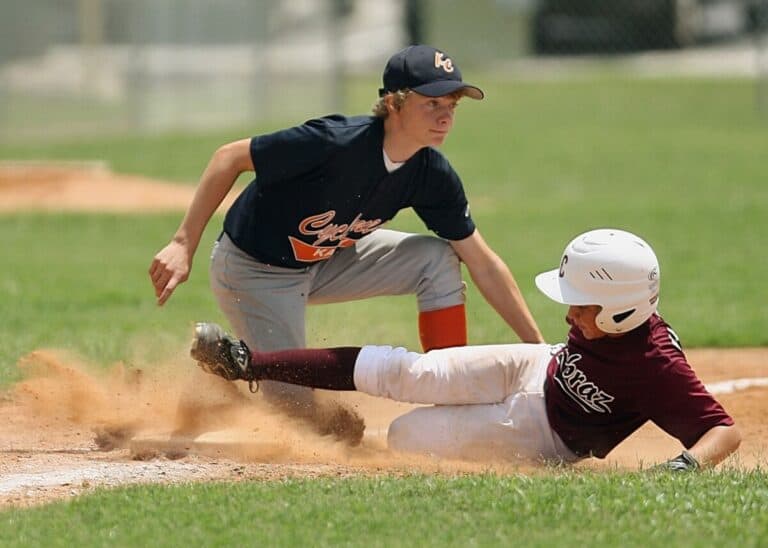 Baseball base runner in maroon jersey sliding feet-first into third base while the third baseman is preparing to catch the throw
